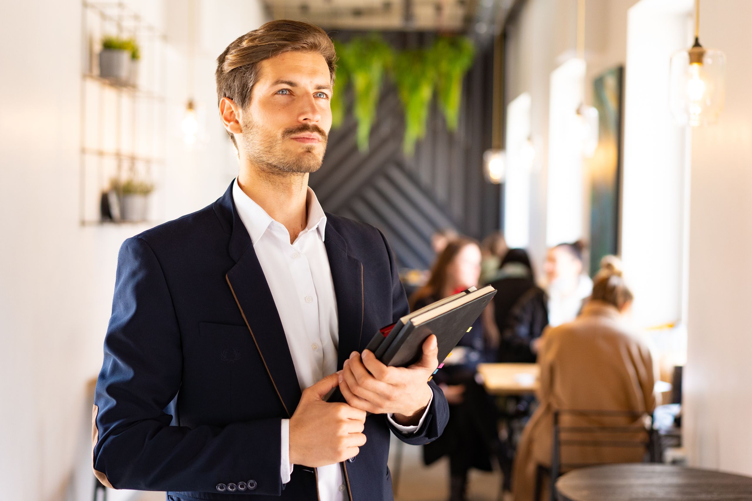 Karriere 1 Confident man in a blue suit holding notebooks in a modern cafe setting.
